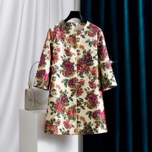 May include: A cream-colored coat featuring a vibrant floral pattern in shades of magenta, deep pink, and dark green. The coat has a round neckline, three-quarter length sleeves, and a straight silhouette, hanging on a black hanger.