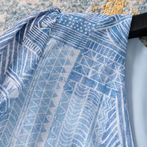 May include: Close-up of a light blue garment with a white geometric pattern. The fabric features a repeating design of lines, triangles, and other shapes. The garment is hanging on a black hanger.
