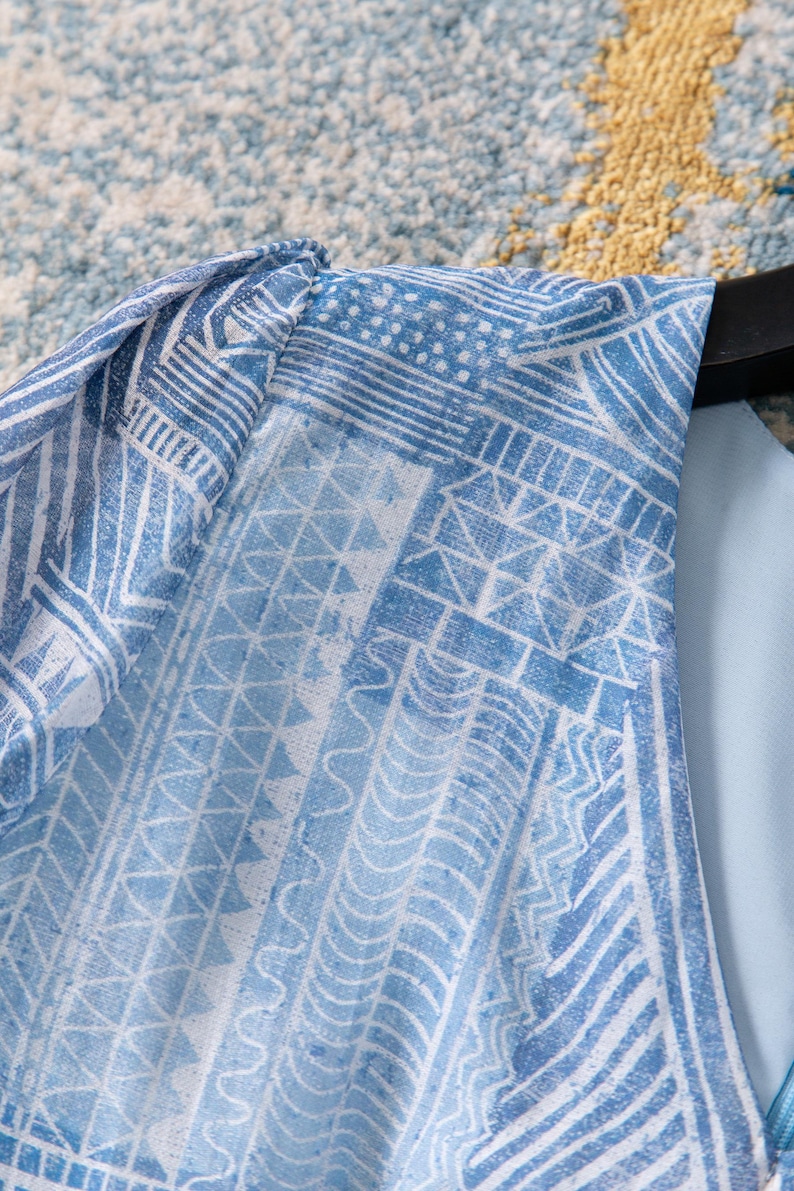May include: Close-up of a light blue garment with a white geometric pattern. The fabric features a repeating design of lines, triangles, and other shapes. The garment is hanging on a black hanger.
