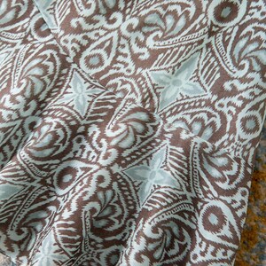 Op de afbeelding: Close-up van een stof met een herhalend paisley-patroon in bruin en lichtblauw. De stof lijkt gedrapeerd te zijn, met plooien en schaduwen die diepte aan het ontwerp toevoegen. Het patroon is gedetailleerd en ingewikkeld.