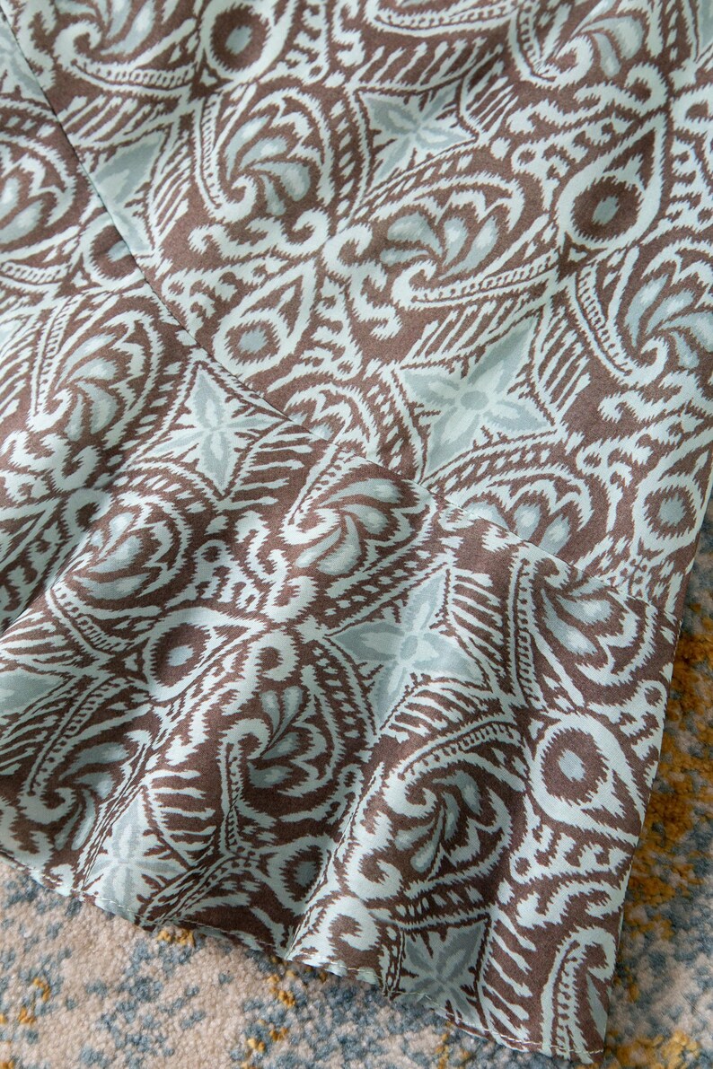 Op de afbeelding: Close-up van een stof met een herhalend paisley-patroon in bruin en lichtblauw. De stof lijkt gedrapeerd te zijn, met plooien en schaduwen die diepte aan het ontwerp toevoegen. Het patroon is gedetailleerd en ingewikkeld.