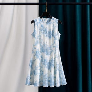 Op de afbeelding: Mouwloze wit en blauwe jurk met een bloemenpatroon. De jurk hangt aan een zwarte hanger. De jurk heeft een ronde halslijn en een uitlopende rok. De stof lijkt lichtgewicht en geschikt voor warm weer.