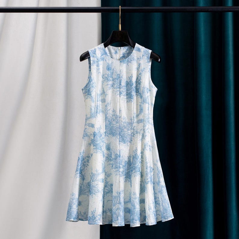 Op de afbeelding: Mouwloze wit en blauwe jurk met een bloemenpatroon. De jurk hangt aan een zwarte hanger. De jurk heeft een ronde halslijn en een uitlopende rok. De stof lijkt lichtgewicht en geschikt voor warm weer.