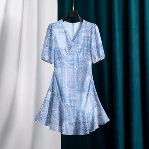 May include: Light blue dress with a V-neck and short sleeves. The dress features a geometric pattern in white and blue. The skirt has a ruffled hem. The dress is hanging on a black hanger.