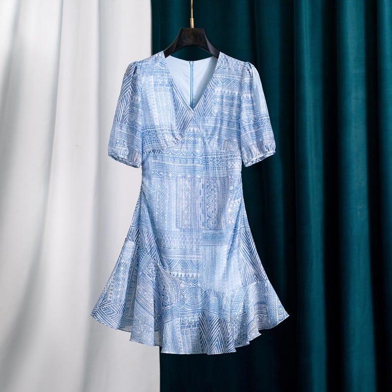 May include: Light blue dress with a V-neck and short sleeves. The dress features a geometric pattern in white and blue. The skirt has a ruffled hem. The dress is hanging on a black hanger.