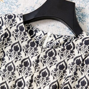 Op de afbeelding: Close-up van een cr&egrave;mekleurig kledingstuk met een zwart bloemmotief. De stof is bedrukt met gestileerde zwarte bloemen en bladeren. Een zwarte hanger ondersteunt het kledingstuk en een witte rits is zichtbaar.