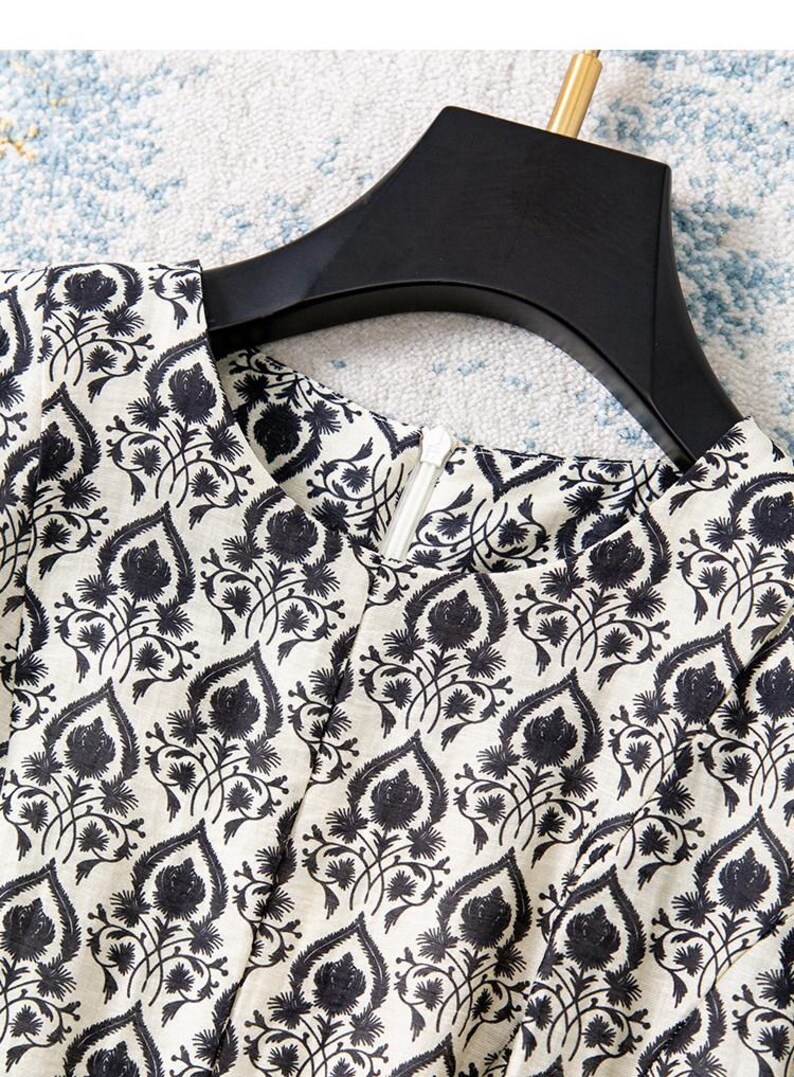 Op de afbeelding: Close-up van een cr&egrave;mekleurig kledingstuk met een zwart bloemmotief. De stof is bedrukt met gestileerde zwarte bloemen en bladeren. Een zwarte hanger ondersteunt het kledingstuk en een witte rits is zichtbaar.