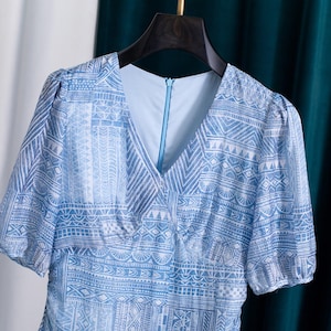 May include: A light blue dress with a V-neck and short sleeves. The dress features a white geometric pattern. The dress is hanging on a black hanger against a white and teal background.