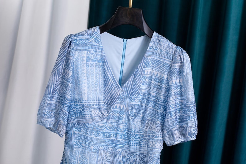 May include: A light blue dress with a V-neck and short sleeves. The dress features a white geometric pattern. The dress is hanging on a black hanger against a white and teal background.