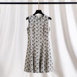 Op de afbeelding: Mouwloze jurk met een zwart-wit bloemenpatroon. De jurk hangt aan een zwarte hanger tegen een witte achtergrond. De jurk heeft een ronde halslijn en een getailleerd silhouet.