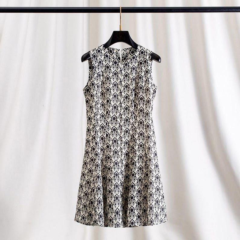 Op de afbeelding: Mouwloze jurk met een zwart-wit bloemenpatroon. De jurk hangt aan een zwarte hanger tegen een witte achtergrond. De jurk heeft een ronde halslijn en een getailleerd silhouet.