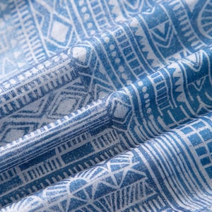 May include: Close-up of a blue and white fabric with a geometric pattern. The design features repeating shapes, lines, and dots, creating a textured appearance. The fabric is folded, showing depth and dimension.