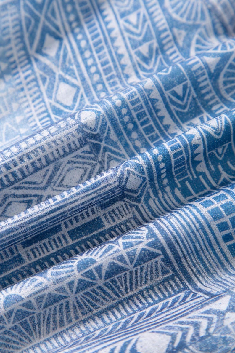May include: Close-up of a blue and white fabric with a geometric pattern. The design features repeating shapes, lines, and dots, creating a textured appearance. The fabric is folded, showing depth and dimension.