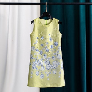 Puede incluir: Vestido sin mangas, hasta la rodilla, en tono amarillo pálido. El vestido presenta un estampado floral en tonos azules y blancos, con sutiles adornos. Cuelga de una percha negra sobre un fondo de cortinas blancas y verde azulado. Mide unos 90 cm.