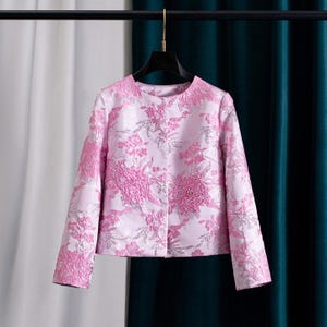 Könnte beinhalten: Eine rosa Jacke mit floralem Muster. Die Jacke hat lange Ärmel und einen runden Ausschnitt. Das florale Design ist in einem dunkleren Rosa- und Silberschattierung gehalten und erzeugt einen strukturierten Effekt. Die Jacke hängt an einem schwarzen Kleiderbügel.