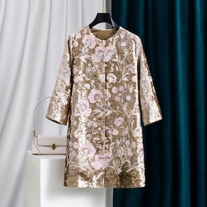 May include: A long-sleeved, knee-length coat in a metallic gold velvet fabric. The coat features a round neckline and is decorated with an all-over floral pattern embroidered in shades of light pink, white, and silver sequins and thread.