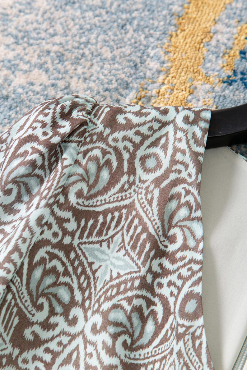 Op de afbeelding: Close-up van een stuk stof met een herhalend paisley-patroon. Het patroon heeft bruine en lichtblauwe wervelingen en bloemvormen op een witte achtergrond. De stof is over een zwarte hanger gedrapeerd.