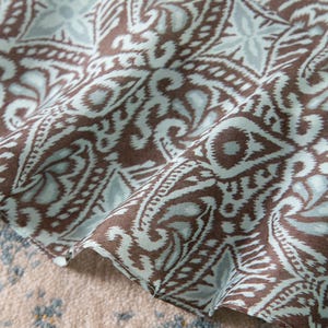Op de afbeelding: Close-up van een stof met een herhalend paisley-patroon in bruin en lichtblauw. De stof is gevouwen en onthult de ingewikkelde details van het patroon. Het ontwerp bevat bloemen en geometrische elementen.