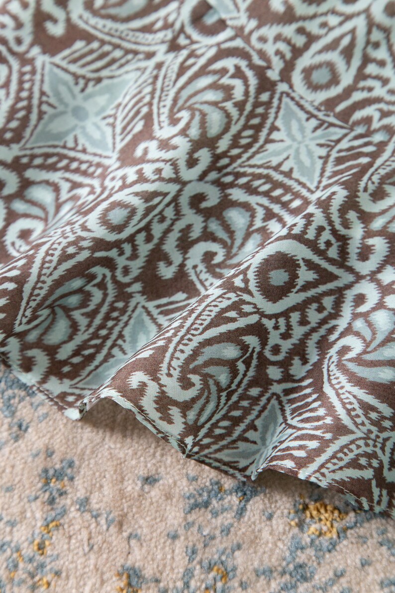 Op de afbeelding: Close-up van een stof met een herhalend paisley-patroon in bruin en lichtblauw. De stof is gevouwen en onthult de ingewikkelde details van het patroon. Het ontwerp bevat bloemen en geometrische elementen.