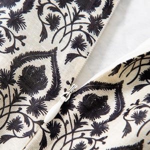 Op de afbeelding: Close-up van een cr&egrave;mekleurige stof met een zwart bloemenpatroon. Het ontwerp toont gestileerde bloemen en bladeren. Een witte rits is gedeeltelijk zichtbaar, wat wijst op een kledingstuk of textielartikel.