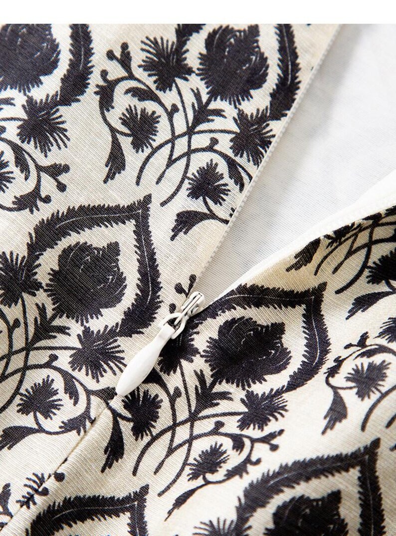 Op de afbeelding: Close-up van een cr&egrave;mekleurige stof met een zwart bloemenpatroon. Het ontwerp toont gestileerde bloemen en bladeren. Een witte rits is gedeeltelijk zichtbaar, wat wijst op een kledingstuk of textielartikel.