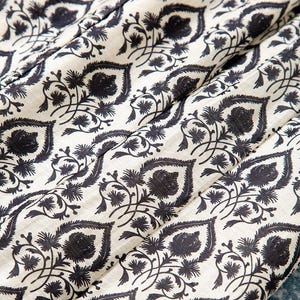 Op de afbeelding: Close-up van een stof met een herhalend patroon van donker marineblauwe bloemen- en bladmotieven op een gebroken witte achtergrond. De stof is gevouwen en toont de textuur en de drapering van het materiaal. Het ontwerp doet denken aan traditionele blokprints.