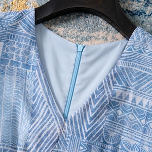 May include: A light blue dress with a V-neckline and a geometric white pattern. The dress has a zipper closure and is hanging on a black hanger. The fabric appears to be lightweight.