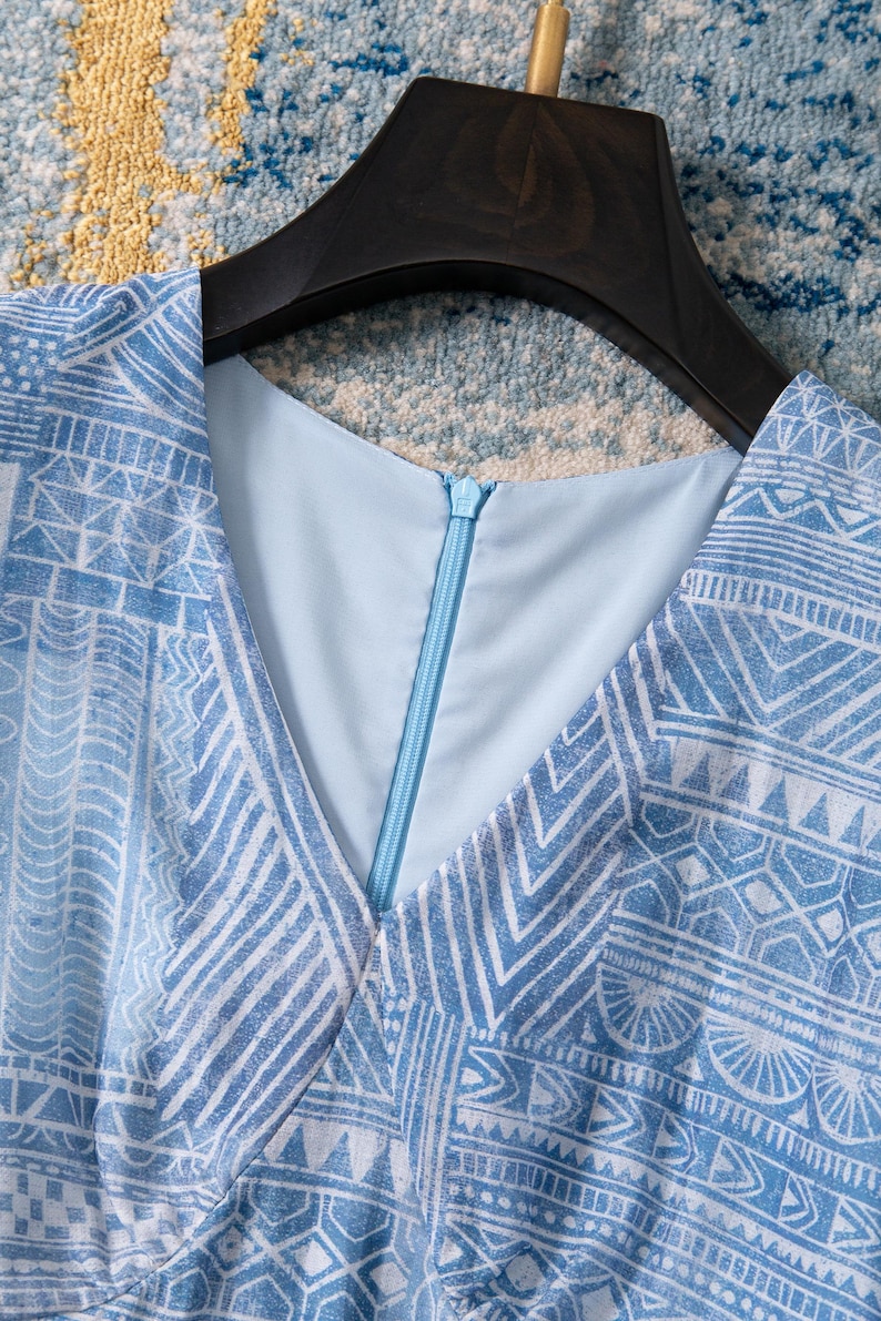 May include: A light blue dress with a V-neckline and a geometric white pattern. The dress has a zipper closure and is hanging on a black hanger. The fabric appears to be lightweight.