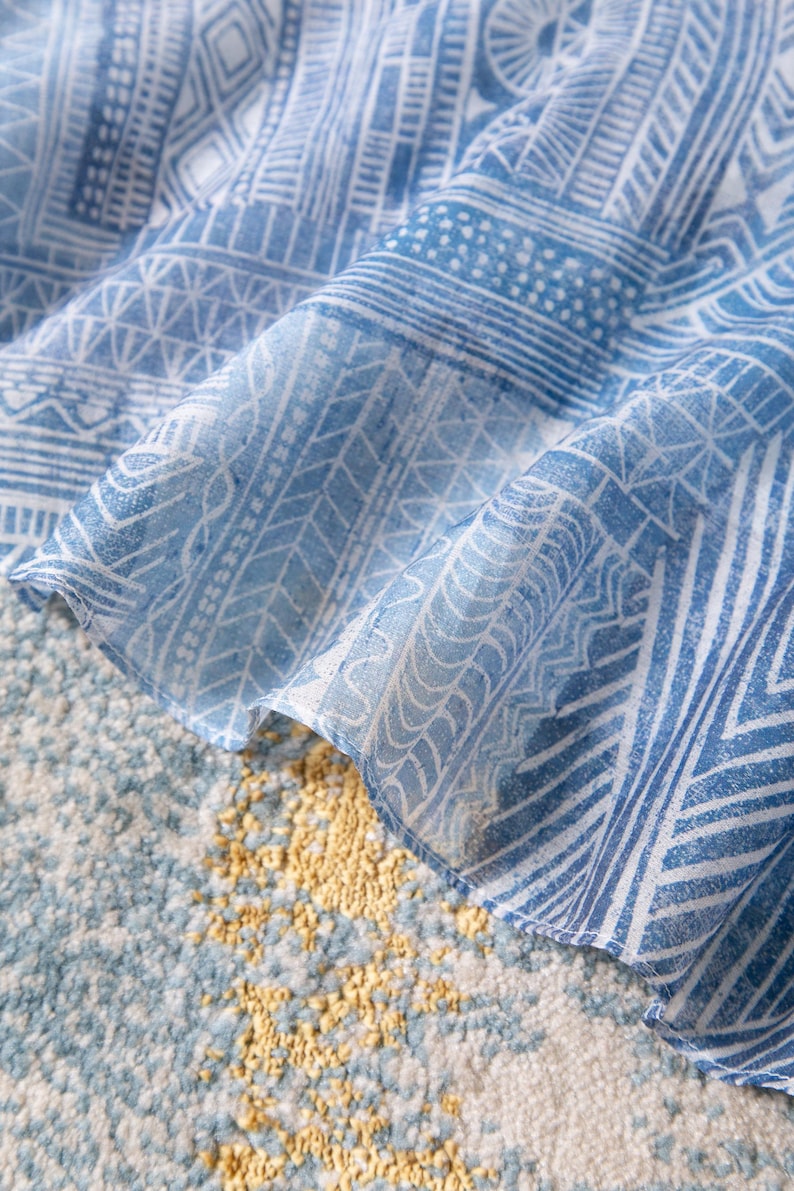 May include: Close-up of a blue and white patterned fabric with geometric designs. The fabric is draped over a textured surface with a mix of blue, white, and yellow tones. The pattern includes lines, shapes, and dotted details.