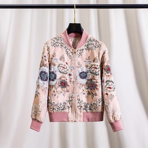 以下が含まれることがあります： フローラル柄のライトピンクのボンバージャケット。 ジャケットはジッパー開閉、ピンクの襟、ピンクの袖口が特徴です。 フローラルデザインには、白、赤、青のアクセントが含まれています。