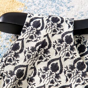 Op de afbeelding: Close-up van een cr&egrave;mekleurige stof met een zwart bloemenpatroon. Het ontwerp bevat gestileerde bladeren en bloemen, waardoor een zich herhalend motief ontstaat. De stof is over een zwarte hanger gedrapeerd.