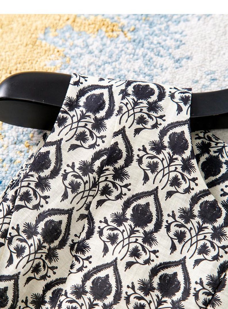 Op de afbeelding: Close-up van een cr&egrave;mekleurige stof met een zwart bloemenpatroon. Het ontwerp bevat gestileerde bladeren en bloemen, waardoor een zich herhalend motief ontstaat. De stof is over een zwarte hanger gedrapeerd.