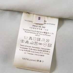 Op de afbeelding: Close-up van een wit kledinglabel met zwarte tekst, inclusief het woord "TROUSERS" en maat "S". Het label bevat ook verzorgingssymbolen en de tekst "100% Lyocell". De achtergrond is een lichtgrijze stof.