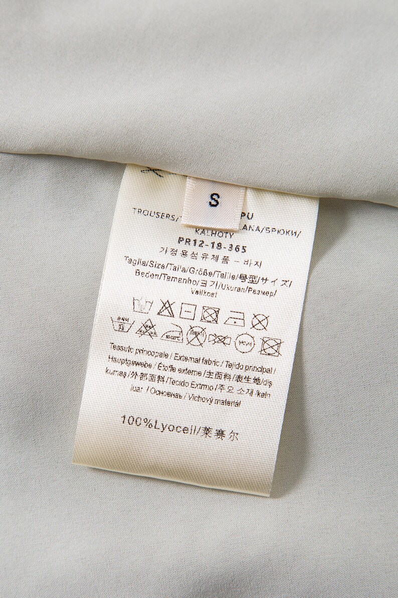 Op de afbeelding: Close-up van een wit kledinglabel met zwarte tekst, inclusief het woord "TROUSERS" en maat "S". Het label bevat ook verzorgingssymbolen en de tekst "100% Lyocell". De achtergrond is een lichtgrijze stof.