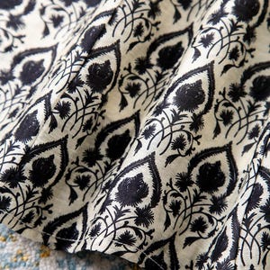 Op de afbeelding: Close-up van een stof met een herhalend zwart bloemenpatroon op een gebroken witte achtergrond. Het ontwerp bevat gestileerde bladeren en bloemelementen, waardoor een gestructureerd, decoratief oppervlak ontstaat. De stof is gevouwen en toont diepte en dimensie.