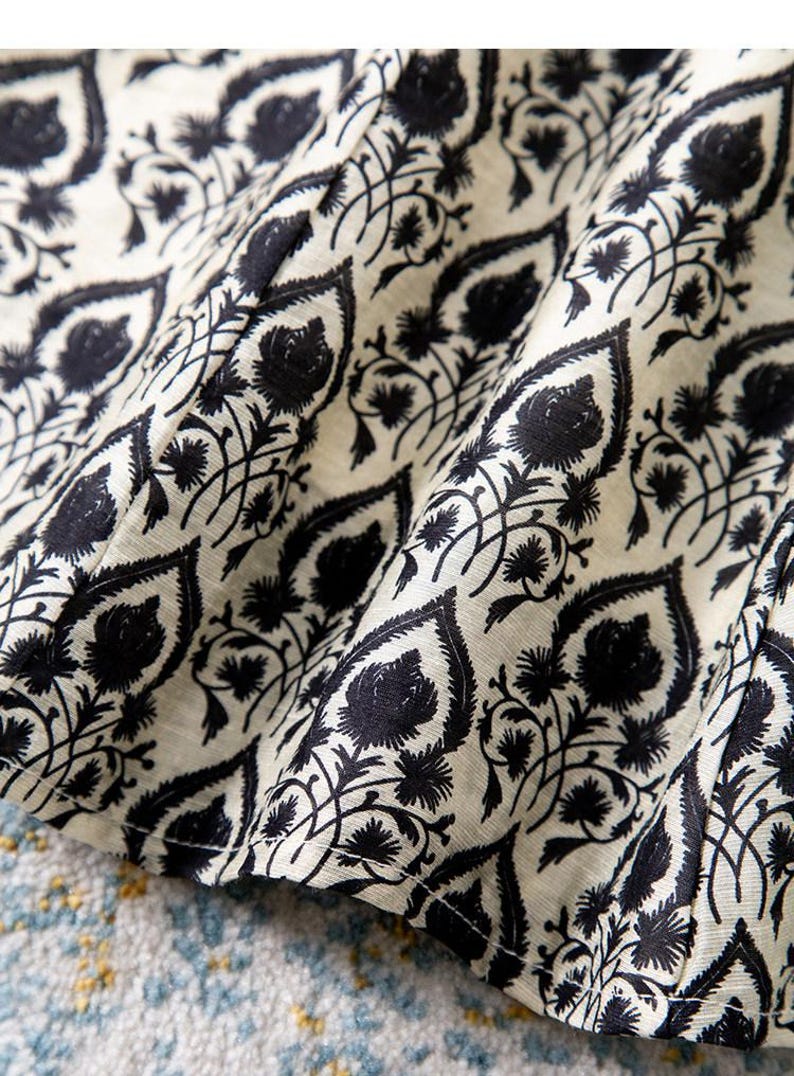Op de afbeelding: Close-up van een stof met een herhalend zwart bloemenpatroon op een gebroken witte achtergrond. Het ontwerp bevat gestileerde bladeren en bloemelementen, waardoor een gestructureerd, decoratief oppervlak ontstaat. De stof is gevouwen en toont diepte en dimensie.