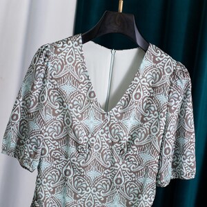 Op de afbeelding: Een jurk met korte mouwen met een V-hals en een bruin, wit en lichtblauw patroon. De jurk hangt aan een zwarte hanger tegen een witte en blauwgroene achtergrond. De stof heeft een bloemen- en geometrisch patroon.