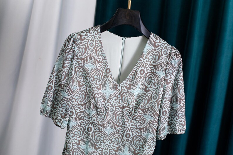 Op de afbeelding: Een jurk met korte mouwen met een V-hals en een bruin, wit en lichtblauw patroon. De jurk hangt aan een zwarte hanger tegen een witte en blauwgroene achtergrond. De stof heeft een bloemen- en geometrisch patroon.