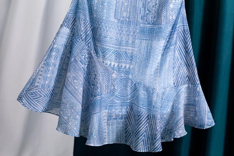 May include: A flowing, light blue skirt with a white geometric pattern. The fabric drapes gracefully, showcasing a detailed design of lines, triangles, and other shapes. The skirt has a ruffled hemline.