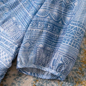 May include: Close-up of a blue and white patterned fabric. The fabric features a repeating geometric design with intricate details. The sleeves of a garment are visible, with a gathered cuff. The fabric is likely a lightweight material.