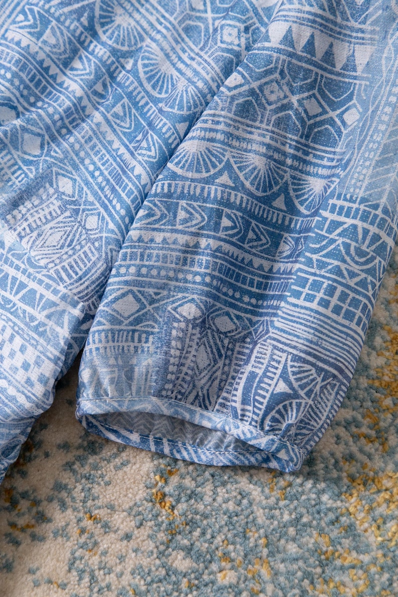 May include: Close-up of a blue and white patterned fabric. The fabric features a repeating geometric design with intricate details. The sleeves of a garment are visible, with a gathered cuff. The fabric is likely a lightweight material.