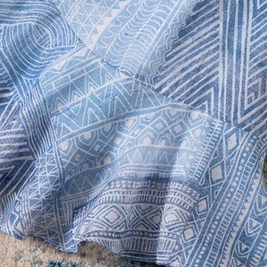 May include: Close-up of a light blue and white patterned fabric. The fabric features a repeating geometric design with lines, triangles, and diamond shapes. The fabric is slightly ruffled, suggesting it may be part of a garment.