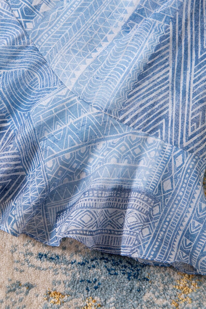 May include: Close-up of a light blue and white patterned fabric. The fabric features a repeating geometric design with lines, triangles, and diamond shapes. The fabric is slightly ruffled, suggesting it may be part of a garment.