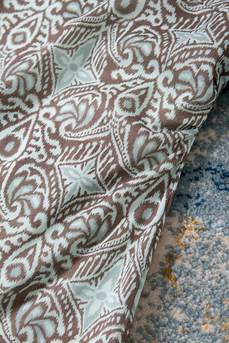 Op de afbeelding: Close-up van een stof met een herhalend patroon in bruin, wit en lichtblauw. Het ontwerp bevat bloem- en geometrische vormen. De stof is gedrapeerd over een blauw en wit patroon tapijt.