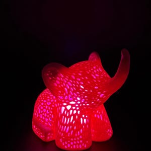 May include: A glowing red elephant-shaped night light. The light has a textured surface with numerous small holes, creating a dotted pattern. The elephant's form is illuminated from within, casting a warm, ambient glow against a black background.