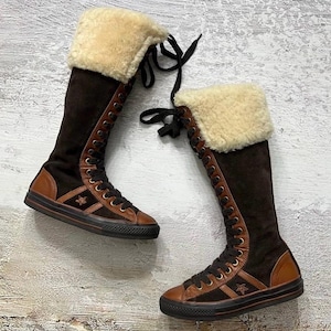 以下が含まれることがあります： 星のロゴが入った茶色とタン色のニーハイブーツ。ブーツはレースアップデザイン、タン色のフェイクファーカフ、黒いソールが特徴です。ブーツはスエードと革で作られています。