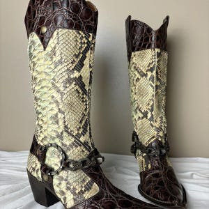 May include: A pair of tall cowboy boots with a pointed toe. The boots feature a snakeskin pattern in cream and brown, with dark brown leather accents and a decorative buckle. The heels are dark brown.
