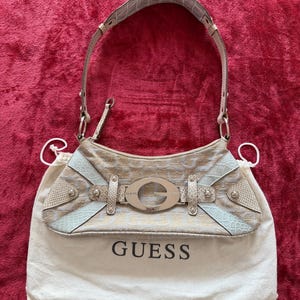 May include: A beige and light blue shoulder bag featuring a repeating gold logo pattern, a large silver-tone metal G logo buckle, and textured faux-reptile skin accents. The bag includes a single handle and rests on a white fabric dust bag with GUESS printed.