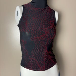 May include: Black sleeveless top with a mock turtleneck. The top features a red, net-like pattern over the front and back. The top is displayed on a mannequin.