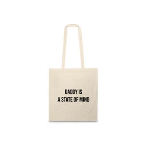 Può includere: Borsa tote color crema con la scritta nera "DADDY IS A STATE OF MIND". La borsa ha due manici ed è realizzata in tessuto resistente. Il design è semplice, con il testo centrato, adatta per trasportare vari oggetti.